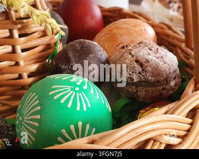 Eine Nahaufnahme eines geflochtenen Osterkorbs mit verzierten Eiern und Süßigkeiten. Im Vordergrund zeigt ein leuchtend grünes Osterei Stockfoto