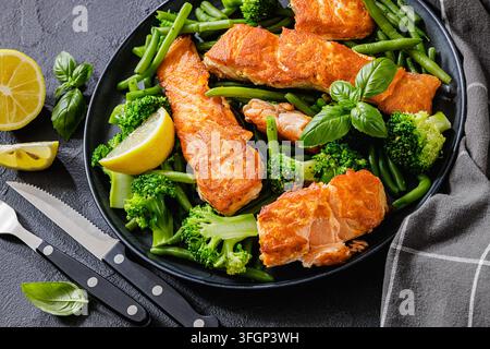 An der Pfanne gebratener Lachs, serviert mit gedämpften grünen Bohnen, Brokkolischöseln, frischen Zitronenspaltern auf einem Teller mit Besteck auf dunklem, texturiertem Tisch, horizontal Stockfoto