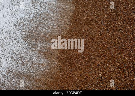 Die Wellen spülen sanft über einen Strand, der mit kleinen Kieselsteinen bedeckt ist. Das Wasser und die Kieselsteine erzeugen Muster, während sie miteinander wechselwirken. Sonnenlicht erhellt die Szene. Stockfoto