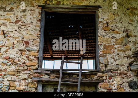 Großes offenes Fenster, eingerahmt von verwittertem Holz und grober Steinmauer, mit einer alten Holzleiter und Ziegeldachkonstruktion, die durch die Öffnung sichtbar ist. Stockfoto