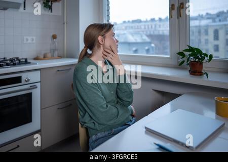 Vom Stress erschöpfte junge Frau sitzt am Tisch und berührt nervös ihr Gesicht, überwältigt von Gedanken in der Nähe eines geschlossenen Laptops. Müde besorgte Frau schaut zum Fenster und fühlt emotionalen Burnout, Frustration zu Hause. Stockfoto
