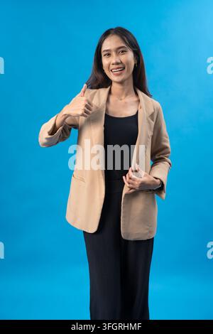 Selbstbewusste junge asiatische Geschäftsfrau in einem beigen Blazer, die ein Daumen-hoch-Zeichen gibt, während sie präsentiert, isoliert vor einem hellblauen Studiohintergrund. Stockfoto