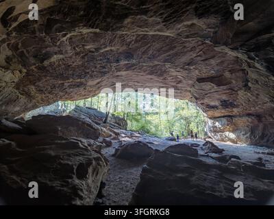 Eine niedrige Blickwinkelansicht aus einem großen natürlichen Felsschutz heraus auf einen sonnenbeschienenen Wald mit Besuchern, die auf dem Pfad spazieren. Stockfoto