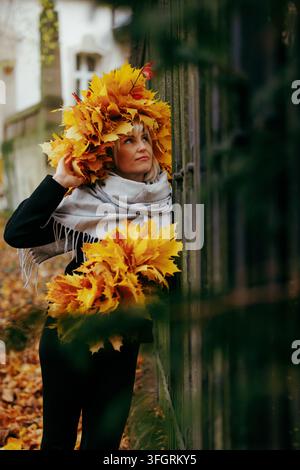 Eine verträumte blonde Frau, die ein großes, kreatives Kopfstück und ein Outfit-Element aus gelben Herbst-Ahornblättern trägt, posiert draußen neben einem Vintage-Messgerät Stockfoto