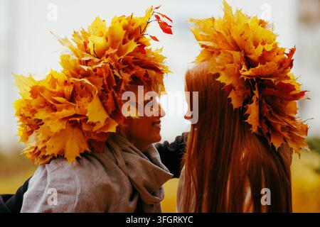 Ein Seitenansicht-Porträt von zwei glücklichen Frauen, eine blonde und eine rothaarige, die große kreative Kränze aus lebendigen gelben Herbst-Ahornblättern tragen Stockfoto