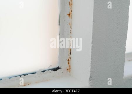Rostflecken und schwarzer Schimmel breiten sich entlang eines rissigen weißen Fensterrahmens mit abblätternder Farbe an einer feuchten Wand aus. Nahaufnahme-Detail für die Hausinspektion, Feuchtigkeit Stockfoto