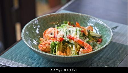 Caesar-Salat mit gegrillten Garnelen, Römersalat, Croutons und Parmesan serviert in einer grünen Schüssel auf dem Restauranttisch. Gesunde Meeresfrüchte-Mahlzeit, Kulinarik Stockfoto