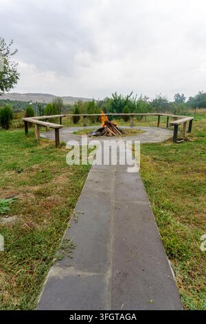Gepflasterter Weg auf Rasen, der zu einer Feuerstelle führt, die von Bänken in einem Kreis umgeben ist. Brennende Holzscheite. Hügel und bewölkter Himmel im Hintergrund Stockfoto