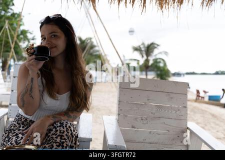 Eine tätowierte junge Frau mit einer Sonnenbrille auf dem Kopf entspannt sich auf einem weißen Holzstrandstuhl und nippt an Kaffee aus einer kleinen schwarzen Tasse unter einem Strohdach Stockfoto
