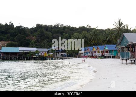 Lebendig bemalte Holzbungalows und Pfahlhäuser säumen einen weißen Sandstrand auf einer tropischen Insel in Südostasien, mit üppigen, dschungelbedeckten Hügeln im Stockfoto