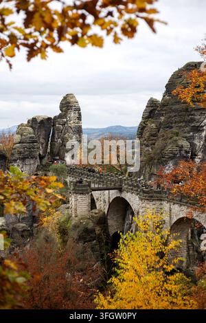 Vertikale Aufnahme der ikonischen Sandstein-Bastei-Brücke mit Touristen, eingerahmt von warmem gelbem und orangefarbenem Herbstlaub in der Sächsischen Schweiz. Stockfoto