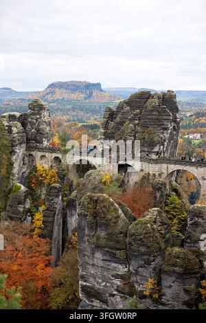 Vertikale Aufnahme, die die Bögen der Bastei-Brücke zeigt, die hohe Sandsteinklippen mit gelbem Herbstwald verbindet, der in tiefen Schluchten darunter wächst. Stockfoto