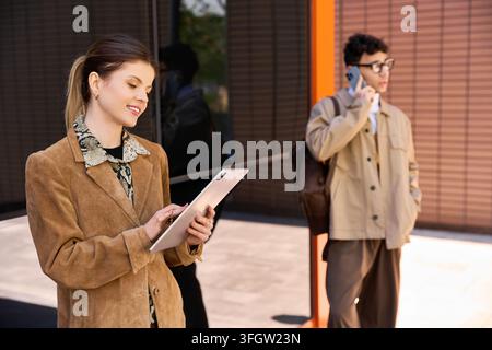 Ein Mann und eine Frau zeigen Professionalität, während sie digitale Geräte außerhalb eines schlanken Gebäudes verwenden. Stockfoto