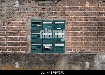 Ein Block aus neun altmodischen grünen Metallbriefkästen mit Namensschlitzen, eingebettet in einer verwitterten roten Ziegelmauer. Stockfoto