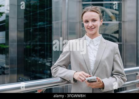 Geschäftsfrau mit Smartphone Stehend lächelnd. Geschäftsfrau kaukasisch weiblich selbstbewusst Porträt Outdoor Stadt. Moderne Unternehmensführung Führungskraft Mobil Co Stockfoto