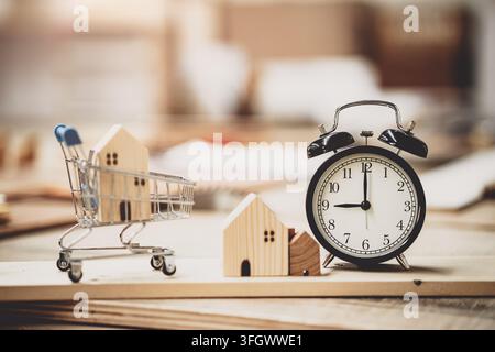 Holzhaus-Modell im Einkaufswagen mit Wecker Zuhause zum Kauf Renovieren Zeitplanung Konzept, Immobilieninvestmentkredit Hypothek Eigentum Fin Stockfoto