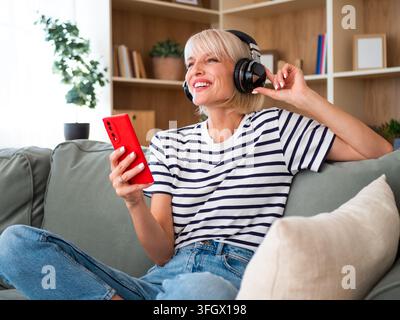Glückliche junge Frau hört ihre Lieblingsplaylist über drahtlose Kopfhörer, während sie ihr Telefon benutzt. Stockfoto