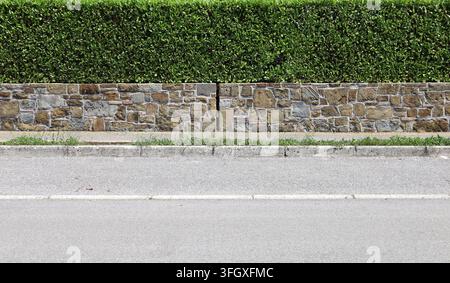Zaun aus Stein mit großen flachen braunen und grauen Natursteinen und Ziegeln. Hohe Hecke dahinter, Gehweg und Straße davor. Hintergrund und Textur. Stockfoto