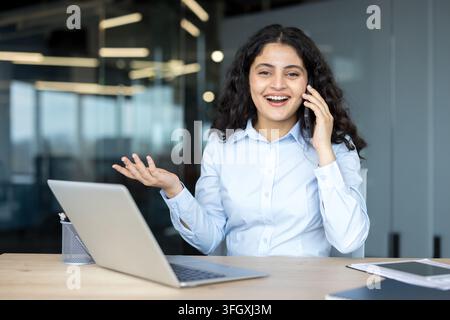 Fröhliche Geschäftsfrau telefoniert am Smartphone und arbeitet am Laptop im Firmenbüro. Professionelle Managerin nutzt Technologie für Kommunikation und Finanzanalyse am Schreibtisch. Selbstbewusste Frau. Stockfoto
