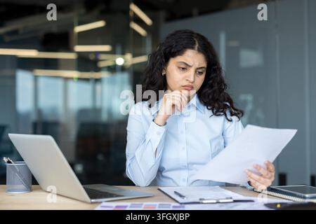 Konzentrierte Geschäftsfrau analysiert Dokumente und Laptop-Daten in einem Firmenbüro. Die professionelle Managerin arbeitet an einer Strategie, während sie wichtige Unterlagen an ihrem Schreibtisch liest. Stockfoto