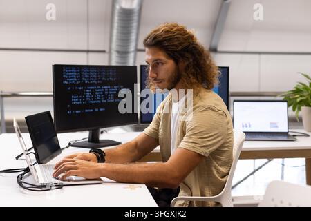 Erwachsener Mann in beigem Overshirt, weißem T-Shirt tippt auf Laptop in einem Open-Space-Büro mit Codemonitor. Programmierer, Codierung, Produktivität, minimalistisch, modern, Stockfoto