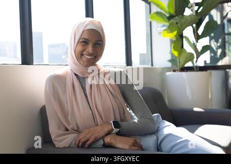 Frau sitzt auf grauem Sofa in moderner Lounge, trägt hellrosa Hijab, überprüft Smartwatch Stockfoto