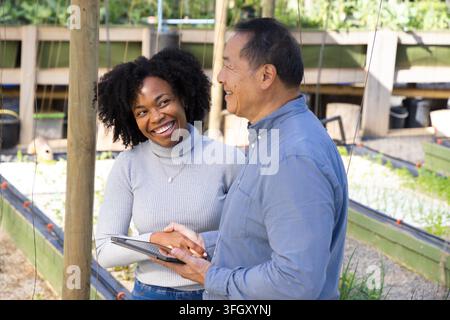 Afroamerikanische Frau und asiatischer Senior arbeiten über einem Tablet im Gewächshaus und inspizieren Sämlinge Stockfoto