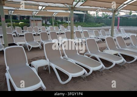 Reihen leerer Plastik-Sonnenliegen mit Seitentischen, die unter einem großen Sonnendach an einem Sandstrand in einem Urlaubsresort platziert sind. Ruhiger Morgen oder Nebensaison Stockfoto