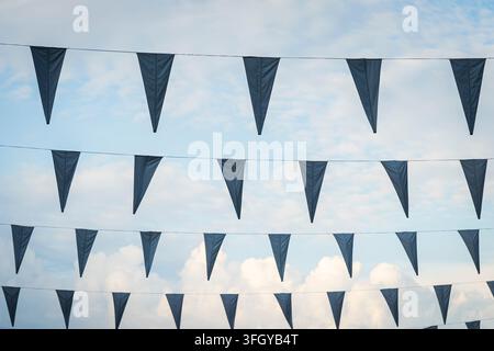 Dreieckige marineblaue Wimpelgirlande, die vor einem hellblauen Himmel mit sanften weißen Wolken hängt und ein klares grafisches Muster aus sich wiederholenden Flaggen erzeugt. Stockfoto