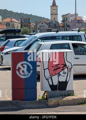 Pirovac, Kroatien, 16. August 2026: Geparkte Autos füllen den Vordergrund neben bunt bemalten Versorgungsboxen, während kleine Küstengebäude und Kirchturm ri Stockfoto