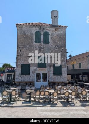 Pirovac, Kroatien, 16. August 2026 Verwitterte Steinfassade mit grünen Fensterläden, kleinem Schornstein und Reihen von Außengastronomie-Stühlen und -Tischen darunter Stockfoto