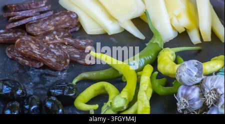 Nahaufnahme von Scheiben gekochter Wurst, Käsestreifen, grünen Paprikaschoten, schwarzen Oliven und Knoblauch, die zusammen auf einem rustikalen Vorspeisenplatte arrangiert sind. Stockfoto