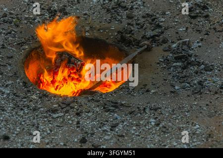 Nahaufnahme von hellen Flammen und glühenden Kohlen in einer kleinen Bodenfeuerstelle mit einem Metallstab, der darin erhitzt wird, was traditionelle Metallverarbeitung im Freien nahelegt Stockfoto
