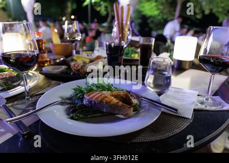 Romantischer Esstisch mit Gläsern rotwein, gegrilltem Fischfilet und moderner Schnurlampe in einem Restaurant im Freien bei Nacht. Stockfoto