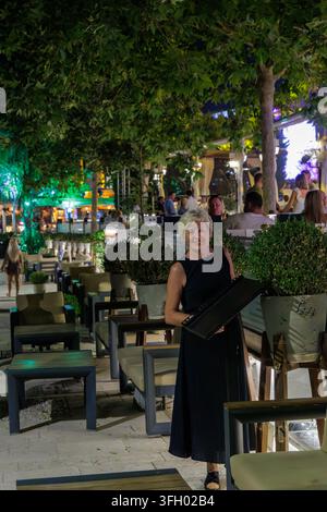 SIDE, TÜRKEI – SEPTEMBER 2026: Fröhliche ältere Dame mit kurzem blondem Haar steht mit einer Speisekarte in einer überfüllten Freiluft-Kaffee-Promenade im Side-Resort. Stockfoto