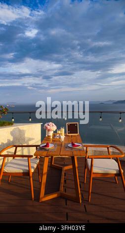 Leerer Holztisch und Stühle auf einem Dachgarten-Café-Balkon mit Blick auf ein ruhiges blaues Meer, eine Küstengebirgslandschaft und grüne tropische Pflanzen unter Stockfoto