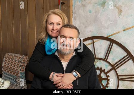 Ein glückliches Paar mittleren Alters, das sich drinnen liebevoll umarmt, wobei eine Frau ihren Ehemann von hinten umarmt, während beide sich warm anlächeln. Stockfoto