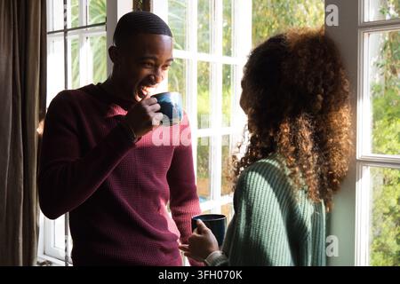 Paar steht in Sweatshirts neben einem geteilten Fenster zu Hause und hält blaue und dunkle Tassen, Sonnenlicht strömt herein Stockfoto
