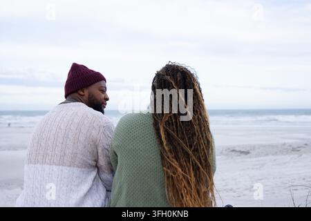 Afrikanisches Paar sitzt auf sandigem Strand mit Blick auf den Ozean, Mann mit brauner Mütze, Frau in grünem Pullover Stockfoto
