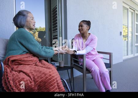 Asiatische Seniorin im Rollstuhl und Betreuerin in rosa Krankenpflegekleidung sitzen auf dem Balkon und halten eine Tasse Stockfoto
