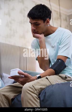 Afroamerikanischer Mann sitzt auf dem Bett im Schlafzimmer, hält einen Monitor und trägt eine Armbinde und ein T-Shirt Stockfoto