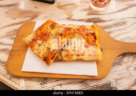 Lipari, ME - IT - 6. Sep 2026 : Zwei rechteckige Scheiben Käse-Tomaten-Pizza, mit Kräutern gewürzt, ruhen auf einem weißen Serviette und einem Holzservierbrett Stockfoto