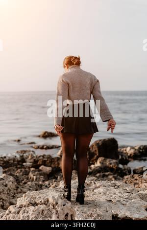 Frau an der Küste steht auf einer felsigen Küstenlinie und blickt während der goldenen Stunde in einem stilvollen Outfit auf den ruhigen Ozean Stockfoto