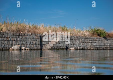 Eine Steinmauer mit eingebauten Treppen steht entlang ruhigen, spiegelnden Wassers, bedeckt mit trockenem Wildgras unter einem klaren Himmel in Secovlje. Stockfoto