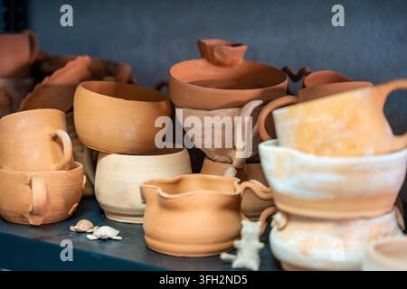 Sammlung handgefertigter Keramikbecher und Schüsseln, die in einer Töpferwerkstatt ausgestellt sind. Unfertige Keramik repräsentiert Handwerkskunst und Stadien der Kunstkeramik Stockfoto