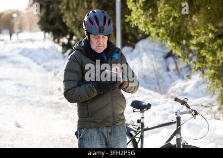Ein älterer Mann überprüft sein Smartphone in der Nähe seines Fahrrads auf einer verschneiten Parkstraße in Helsinki, Finnland, und plant seine Winterpendelreise Stockfoto