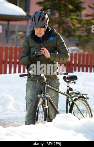 Ein älterer Mann überprüft sein Smartphone in der Nähe seines Fahrrads auf einer verschneiten Parkstraße in Helsinki, Finnland, und plant seine Winterpendelreise Stockfoto