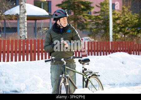 Ein älterer Mann überprüft sein Smartphone in der Nähe seines Fahrrads auf einer verschneiten Parkstraße in Helsinki, Finnland, und plant seine Winterpendelreise Stockfoto