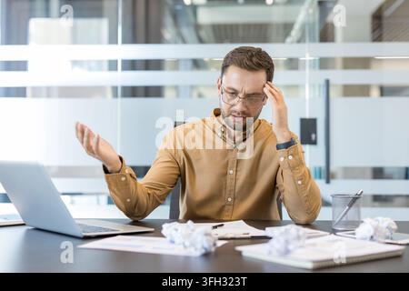 Arbeitsplatz-Burnout und beruflicher Stress, frustrierter Geschäftsmann arbeitet mit Laptop unter Fristdruck und Papierkram im modernen Büro. Erschöpfter Angestellter kämpft mit hoher Arbeitsbelastung. Stockfoto
