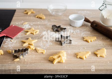 Rohe Weihnachtsplätzchenteig, der mit metallenen festlichen Ausstechformen in Sterne und Bäume geformt wird, mit Nudelholz und Mehl auf einem hölzernen Küchentisch. Stockfoto